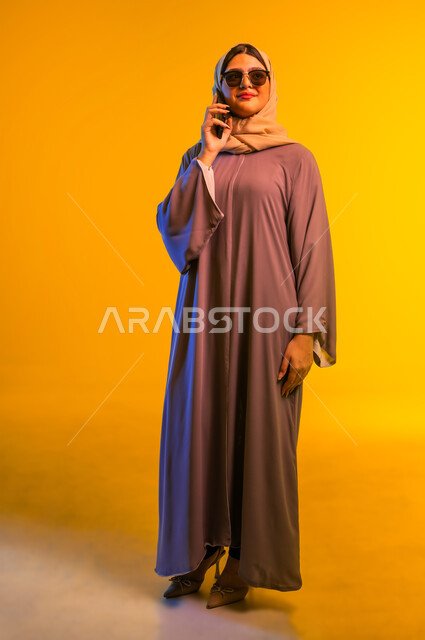 الإجابة والرد على الاتصالات بإستخدام الهاتف المحمول، قضاء اوقات ممتعة بتبادل الاخبار والأحاديث، بورتريه مقرب لامرأة عربية  خليجية عمانية ترتدي عبائة ملونة ونظارة شمسية، شابة سعودية تقوم بإجراء مكالمة صوتية عبر الجوال، التواصل والاطمئنان على الأصدقاء، خلفية صفراء