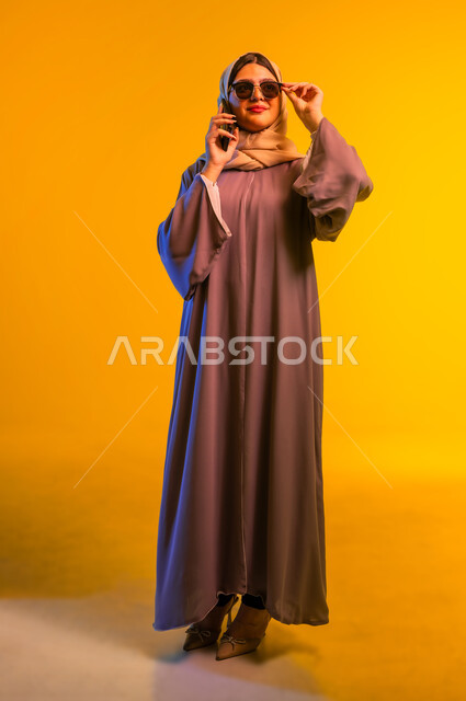 الإجابة والرد على الاتصالات بإستخدام الهاتف المحمول، قضاء اوقات ممتعة بتبادل الاخبار والأحاديث، بورتريه مقرب لامرأة عربية  خليجية عمانية ترتدي عبائة ملونة ونظارة شمسية، شابة سعودية تقوم بإجراء مكالمة صوتية عبر الجوال، التواصل والاطمئنان على الأصدقاء، خلفية صفراء