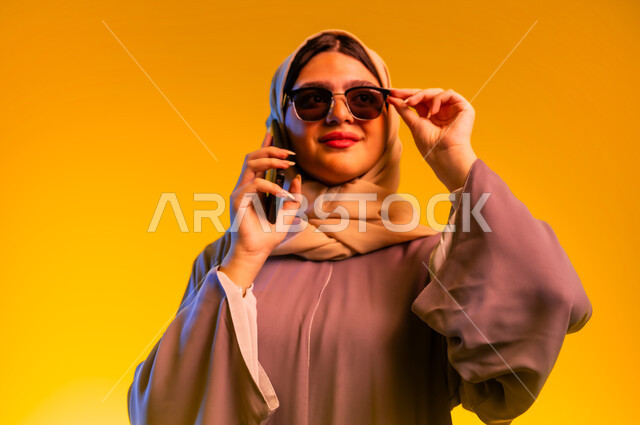 قضاء اوقات ممتعة بتبادل الاخبار والأحاديث، الإجابة والرد على الاتصالات بإستخدام الهاتف المحمول، بورتريه مقرب لامرأة عربية  خليجية عمانية ترتدي عبائة ملونة ونظارة شمسية، شابة سعودية تقوم بإجراء مكالمة صوتية عبر الجوال، التواصل والاطمئنان على الأصدقاء، خلفية صفراء