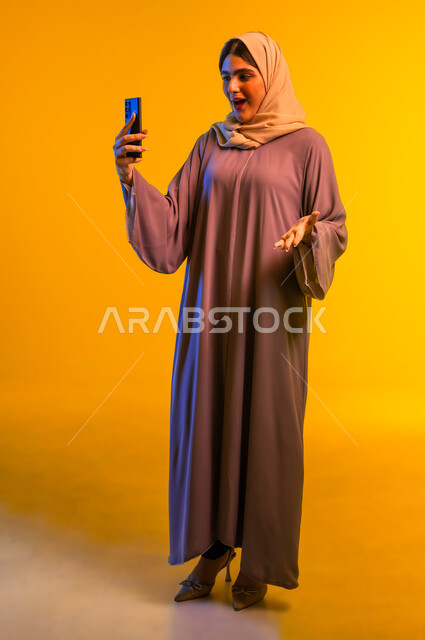 Gestures of surprise and amazement, amazement, wonder and enthusiasm, a young Saudi woman enjoying watching her favorite entertainment programs, close-up portrait of a young Gulf Arab Omani woman wearing a colorful abaya and hijab using mobile phone applications, full-length body, yellow background