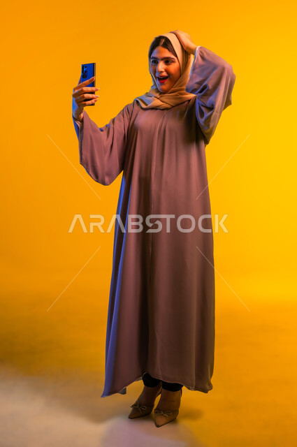 Gestures of surprise and amazement, amazement, wonder and enthusiasm, a young Saudi woman enjoying watching her favorite entertainment programs, close-up portrait of a young Gulf Arab Omani woman wearing a colorful abaya and hijab using mobile phone applications, full-length body, yellow background