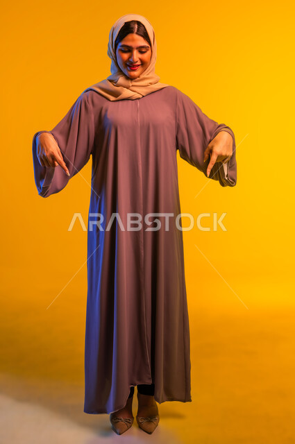 شابة سعودية تحدد موقع شيء معين، إشارات وحركات يدين تدل على هذا المكان (هنا)، بورتريه لامرأة عربية خليجية عمانية محجبة ترتدي العباءة تشير بإصبعي السبابة إلى جهة الأسفل، صورة الجسم بالطول الكامل، خلفية صفراء