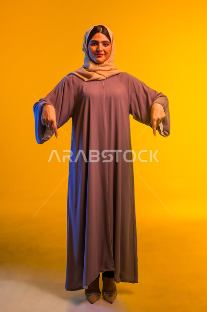 شابة سعودية تحدد موقع شيء معين، إشارات وحركات يدين تدل على هذا المكان (هنا)، بورتريه لامرأة عربية خليجية عمانية محجبة ترتدي العباءة تشير بإصبعي السبابة إلى جهة الأسفل، صورة الجسم بالطول الكامل، خلفية صفراء