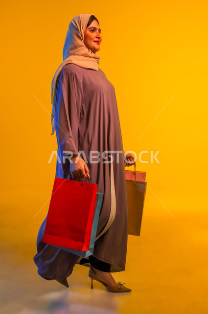 ايماءات المشي والتجول، الشعور بمتعة التسوق، شابة سعودية تستفيد من العروض والتخفيضات، بورتريه لإمرأة عربيه خليجية عمانية ترتدي العباءه تحمل بيدها أكياس التسوق, الاُثر الايجابي للتسوق على نفسية النساء، شراء الهدايا والمستلزمات، الجسم بالطول الكامل، خلفية صفراء