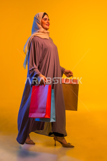 Walking and strolling gestures, feeling the pleasure of shopping, taking advantage of offers and discounts, portrait of an Arab Gulf Omani woman wearing an abaya and hijab holding shopping bags, the positive effect of shopping on women's psychology, buying gifts and supplies, full-length body, yellow background