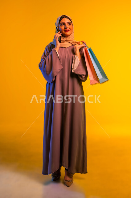 إجراء مكالمة هاتفية, الاستفادة من العروض والتخفيضات الموسمية, استخدام التكنولوجيا والتقنية المتطورة, بورتريه لإمرأة عربية خليجية عمانية ترتدي العباءة تحمل بيدها الأكياس الورقية الملونة، شابة سعودية تجري اتصال عبر الهاتف المحمول, الجسم بالطول الكامل، خلفية صفراء