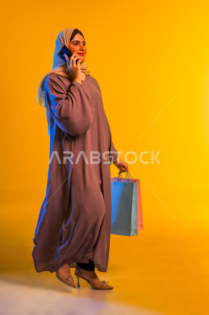 Making a phone call, taking advantage of seasonal offers and discounts, using technology and advanced technology, portrait of an Arab Gulf Omani woman wearing a hijab and abaya holding colorful paper bags in her hand making a call via mobile phone, full body, yellow background