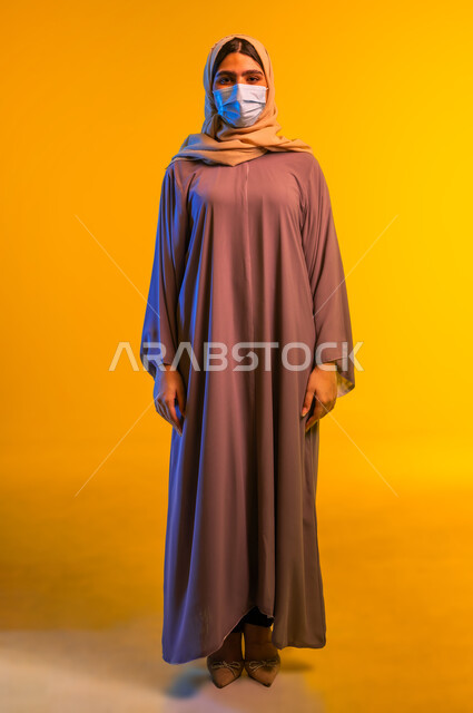 الحفاظ على السلامة من الأمراض والفايروسات, اتباع الإجراءات الاحترازية للوقاية من فايروس كورونا, بورتريه مقرب لامرأة عربية خليجية عمانية محجبة ترتدي العباءة و تضع الكمامة تقف باستقامة، شابة سعودية تنظر الى الكاميرا, الجسم بالطول الكامل، خلفية صفراء