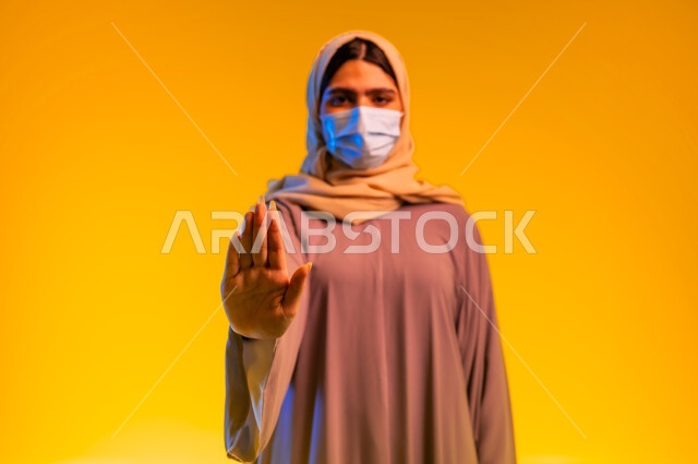 شابة سعودية تتبع الاجراءات الاحترازية والوقائية ضد كوفيد 19, الحفاظ على المسافات وعدم الملامسة السطحية, بورتريه لامرأة عربية خليجية عمانية ترتدي العباءة وتضع الكمامة ترفع كف يدها بتعابير التوقف والابتعاد وتجنب المصافحة, خلفية صفراء