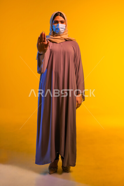 الحفاظ على المسافات وعدم الملامسة السطحية, بورتريه لامرأة عربية خليجية عمانية ترتدي العباءة وتضع الكمامة ترفع كف يدها بتعابير التوقف والابتعاد وتجنب المصافحة, شابة سعودية تتبع الاجراءات الاحترازية والوقائية ضد كوفيد 19, صورة الجسم بالطول الكامل، خلفية صفراء