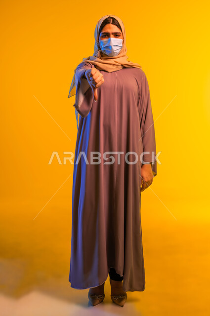 تجنب انتشار العدوى من كوفيد 19, بورتريه لامرأة عربية خليجية عمانية محجبة ترتدي العباءة والكمامة تشير بإصبع الإبهام إلى الأسفل بتعابير عدم الإعجاب والموافقة, شابة سعودية تتبع الإجراءات الاحترازية للوقاية من فايروس كورونا, صورة الجسم بالطول الكامل، خلفية صفراء