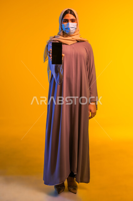 الحفاظ على السلامة من الأمراض والفيروسات, عرض شاشة سوداء فارغة عبر الهاتف المحمول، اتباع الإجراءات الاحترازية للوقاية من فايروس كورونا, بورتريه مقرب لامرأة عربية خليجية عمانية ترتدي العباءة، شابة سعودية تضع الكمامة، الجسم بالطول الكامل، خلفية صفراء