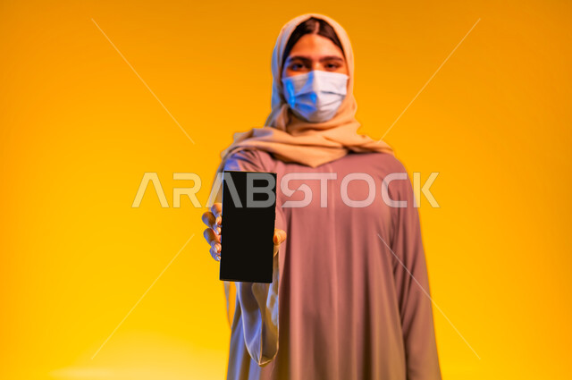 عرض شاشة سوداء فارغة عبر الهاتف المحمول، الحفاظ على السلامة من الأمراض والفيروسات, اتباع الإجراءات الاحترازية للوقاية من فايروس كورونا, بورتريه مقرب لامرأة عربية خليجية عمانية ترتدي العباءة و تضع الكمامة تقف باستقامة، شابة سعودية تنظر الى الكاميرا, خلفية صفراء