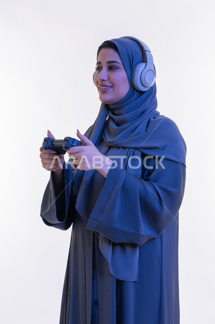 Using modern and advanced electronic technology, close-up portrait from the side of an Arab Gulf Emirati woman wearing an abaya and headphones holding a joystick in her hand, a young Saudi woman playing recreational video games on the weekend, gestures of integration and interaction during the game, white background