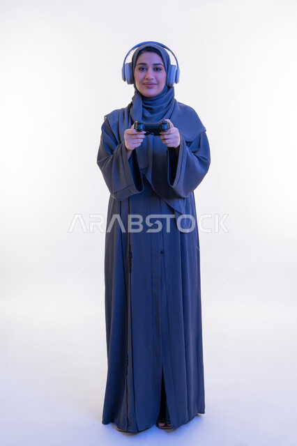 النظر الى الكاميرا بتعابير الفرح والسرور،  قضاء اوقات ممتعة بألعاب الفيديو الترفيهية في عطلة نهاية الاسبوع، بورتريه لإمرأة عربية خليجية إماراتية ترتدي العباءة وسماعة الرأس تحمل بيدها عصا التحكم، شابة سعودية تستخدم التقنية الحديثة، الجسم الكامل بالطول، خلفية بيضاء