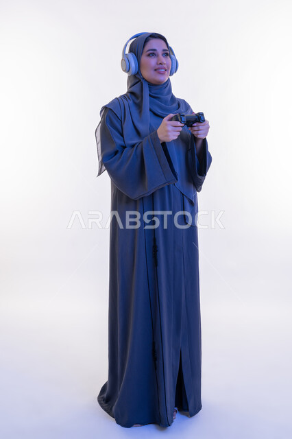 قضاء اوقات ممتعة  بألعاب الفيديو الترفيهية في عطلة نهاية الاسبوع، بورتريه لإمرأة عربية خليجية إماراتية ترتدي العباءة وسماعة الرأس تحمل بيدها عصا التحكم، شابة سعودية تستخدم التقنية الحديثة، ايماءات الاندماج والتفاعل أثناء اللعب، الطول الكامل للجسم، خلفية بيضاء