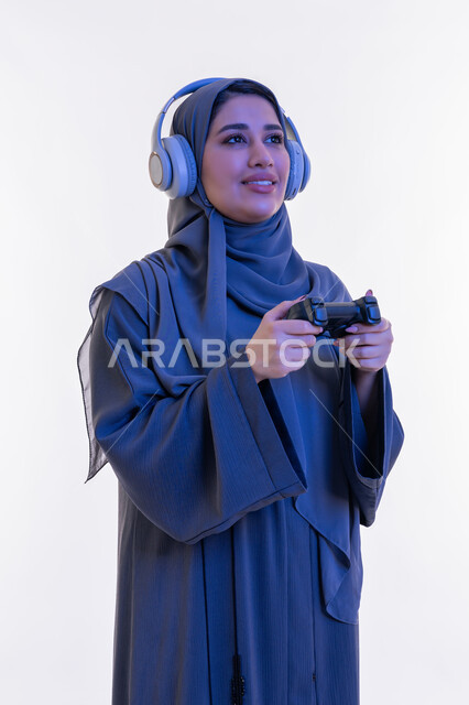 ايماءات الاندماج والاستمتاع أثناء اللعب، قضاء اوقات ممتعة  بألعاب الفيديو الترفيهية في عطلة نهاية الاسبوع، بورتريه مقرب لإمرأة عربية خليجية إماراتية ترتدي العباءة وسماعة الرأس تحمل بيدها عصا التحكم، شابة سعودية تستخدم التقنية الحديثة، خلفية بيضاء