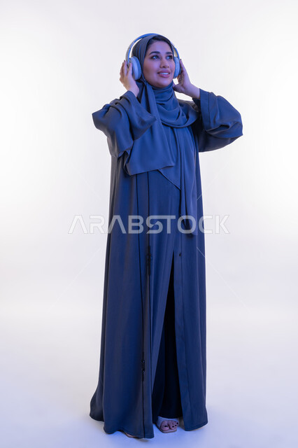 مفهوم الإصغاء والإستماع، قضاء اوقات ممتعة في عطلة نهاية الاسبوع، شابة سعودية تستخدم تقنية الهيدفون، بورتريه  لامرأة عربية خليجية اماراتية محجبة ترتدي العباءة تضع سماعة الرأس الصوتية تستمع إلى الموسيقى بتعابير السعادة والسرور، الجسم الكامل بالطول، خلفية بيضاء