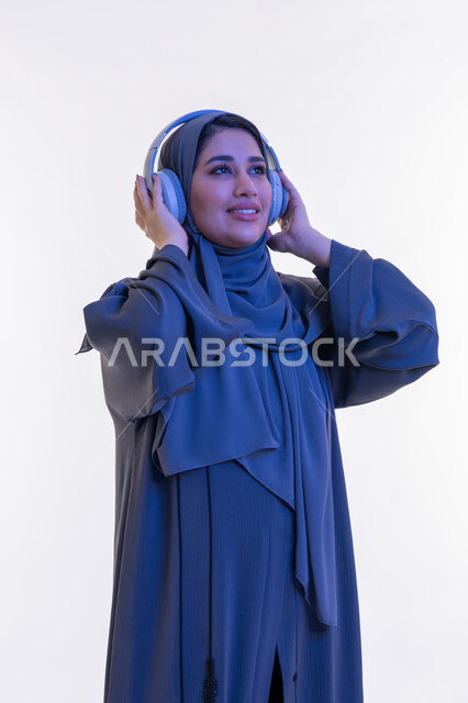قضاء اوقات ممتعة في المنزل في عطلة نهاية الاسبوع، شابة سعودية تستخدم تقنية الهيدفون، مفهوم الإصغاء والإستماع، بورتريه مقرب  لامرأة عربية خليجية اماراتية محجبة مبتسمة ترتدي العباءة تضع سماعات الرأس الصوتية تستمع إلى الموسيقى بتعابير السعادة والسرور، خلفية بيضاء