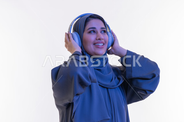 قضاء اوقات ممتعة في المنزل في عطلة نهاية الاسبوع، شابة سعودية تستخدم تقنية الهيدفون، مفهوم الإصغاء والإستماع، بورتريه مقرب  لامرأة عربية خليجية اماراتية محجبة مبتسمة ترتدي العباءة تضع سماعات الرأس الصوتية تستمع إلى الموسيقى بتعابير السعادة والسرور، خلفية بيضاء