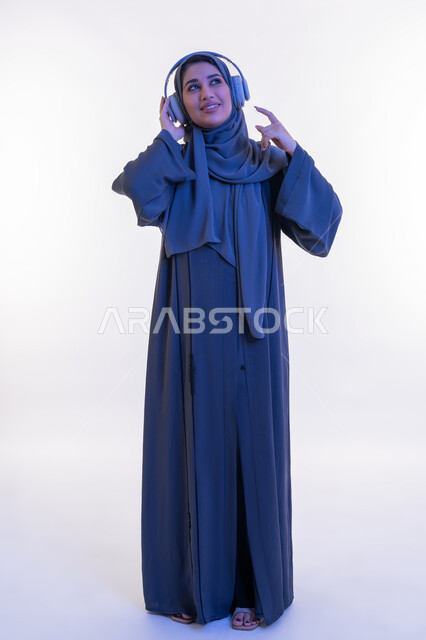 مفهوم الإصغاء والإستماع، قضاء اوقات ممتعة في عطلة نهاية الاسبوع، شابة سعودية تستخدم تقنية الهيدفون، بورتريه  لامرأة عربية خليجية اماراتية محجبة ترتدي العباءة تضع سماعات الرأس الصوتية تستمع إلى الموسيقى بتعابير السعادة والسرور، الجسم الكامل بالطول، خلفية بيضاء