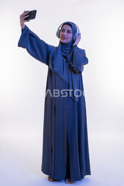 دمج التكنولوجيا في الحياة اليومية، قضاء اوقات ممتعة في عطلة نهاية الاسبوع، شابة سعودية تستخدم تقنية الهيدفون، بورتريه لامرأة عربية خليجية اماراتية محجبة ترتدي العباءة تضع سماعة الرأس تقوم بإلتقاط صور سيلفي باستخدام الجوال، الجسم الكامل بالطول، خلفية بيضاء