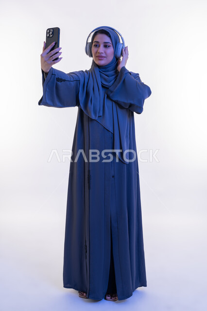 دمج التكنولوجيا في الحياة اليومية، قضاء اوقات ممتعة في عطلة نهاية الاسبوع، شابة سعوية تستخدم تقنية الهيدفون، بورتريه لامرأة عربية خليجية اماراتية محجبة ترتدي العباءة تضع سماعة الرأس الصوتية تقوم بإجراء مكالمة فيديو باستخدام الجوال، الجسم الكامل بالطول، خلفية بيضاء