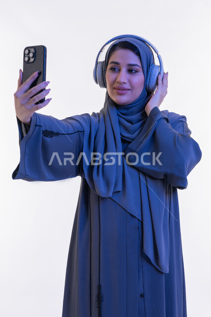 قضاء اوقات ممتعة في عطلة نهاية الاسبوع، شابة سعوية تستخدم تقنية الهيدفون، بورتريه لامرأة عربية خليجية اماراتية محجبة ترتدي العباءة تضع سماعة الرأس الصوتية تقوم بإجراء مكالمة فيديو باستخدام الجوال، دمج التكنولوجيا في الحياة اليومية، خلفية بيضاء