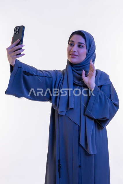 إلقاء التحية والترحيب والسلام، شابة سعودية تستخدم الهاتف المحمول، بورتريه لامرأة عربية خليجية اماراتية ترتدي العباءة تقوم بإجراء مكالمة فيديو عبر تطبيقات التواصل الاجتماعي، التواصل مع الأهل والأصدقاء عبر الانترنت، خلفية بيضاء