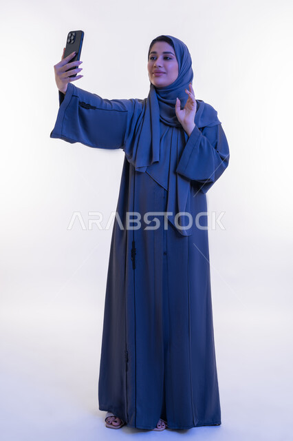 إلقاء التحية والترحيب والسلام، شابة سعودية تستخدم الهاتف المحمول، بورتريه لامرأة عربية خليجية اماراتية ترتدي العباءة تقوم بإجراء مكالمة فيديو عبر تطبيقات التواصل الاجتماعي، التواصل مع الأهل والأصدقاء عبر الانترنت ، الجسم الكامل بالطول، خلفية بيضاء