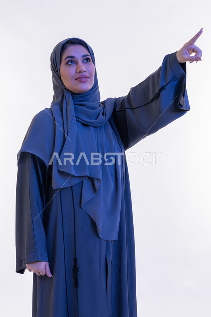 مفهوم النعومة والأنوثة، الاناقة و الاهتمام بالمظهر الخارجي, بورتريه مقرب لامرأة عربيه خليجية إماراتية محجبة ترتدي العباءه  تشير بأصبع السبابة الى مكان ما، شابة سعودية تنظر الى الاعلى بتعابير الفخر والسرور, أزياء محجبات عصرية، خلفية بيضاء