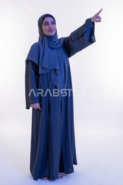 The concept of softness and femininity, elegance and interest in external appearance, portrait of a veiled Arab Gulf Emirati woman wearing an abaya pointing with her index finger to a place, a young Saudi woman looking up with expressions of pride and pleasure, modern veiled fashion, full body image, white background