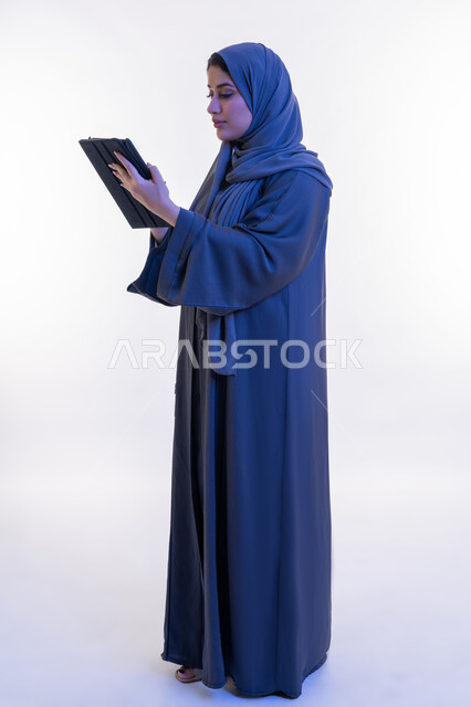 Integrating technology into daily life and work, side portrait of an Arab Gulf Emirati woman wearing an abaya typing on a tablet, a young Saudi woman using an iPad, browsing social media sites, the concept of completing projects remotely, full body length, white background