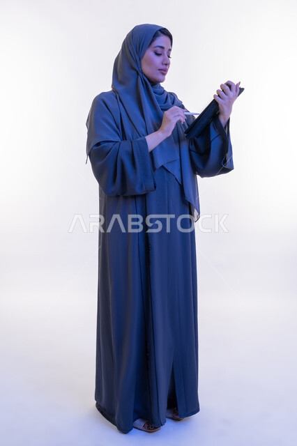 دمج التقنية في الحياة اليومية والعملية، بورتريه من الجانب لامرأة عربية خليجية اماراتية ترتدي العباءة تقوم بالكتابة على جهاز التابلت، شابة سعودية تستخدم الايباد، تصفح مواقع ووسائل التواصل الاجتماعي، مفهوم انجار المشاريع عن بعد، الجسم الكامل بالطول، خلفية بيضاء