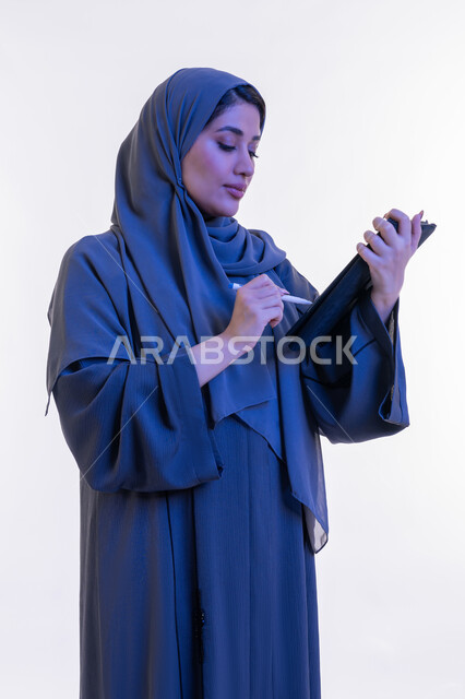 دمج التقنية في الحياة اليومية والعملية، بورتريه مقرب من الجانب لامرأة عربية خليجية اماراتية ترتدي العباءة تقوم بالكتابة على جهاز التابلت، شابة سعودية تستخدم الايباد، تصفح مواقع ووسائل التواصل الاجتماعي، مفهوم انجار المشاريع عن بعد، خلفية بيضاء