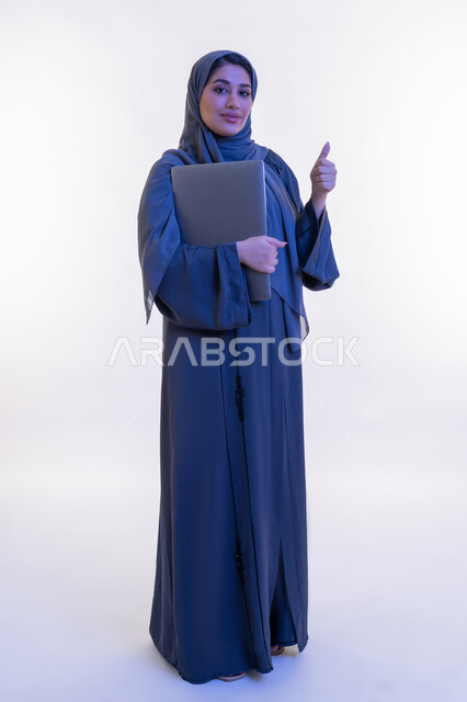 Looking at the camera with expressions of self-confidence and pleasure, a young Saudi woman raising her thumb up in gestures of admiration and approval, integrating modern technology into practical life, portrait of an Arab Gulf Emirati woman wearing an abaya holding a laptop in her hand, full body length, white background