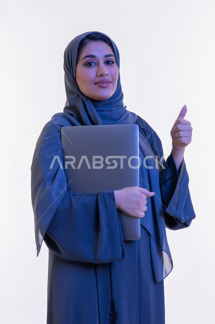 النظر الى الكاميرا بتعابير الثقة بالنفس والسرور، شابة سعودية ترفع اصبع الابهام لاعلى بإيماءات الاعجاب والموافقة, دمج التقنية والتكنولوجيا الحديثة في الحياة العملية، بورتريه مقرب لامرأة عربية خليجية اماراتية ترتدي العباءة  تحمل بيدها جهاز اللابتوب، خلفية بيضاء
