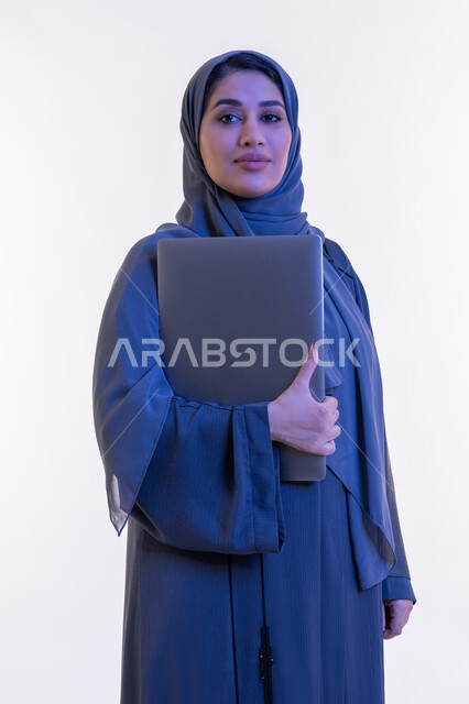 مهن ووظائف نسائية, دمج التقنية والتكنولوجيا الحديثة في الحياة العملية، بورتريه مقرب لامرأة عربية خليجية اماراتية ترتدي العباءة تحمل بيدها جهاز الحاسوب المحمول، شابة سعودية تنظر الى الكاميرا بتعابير الثقة بالنفس والسرور، مفهوم ادارة المشاريع عن بعد، خلفية بيضاء