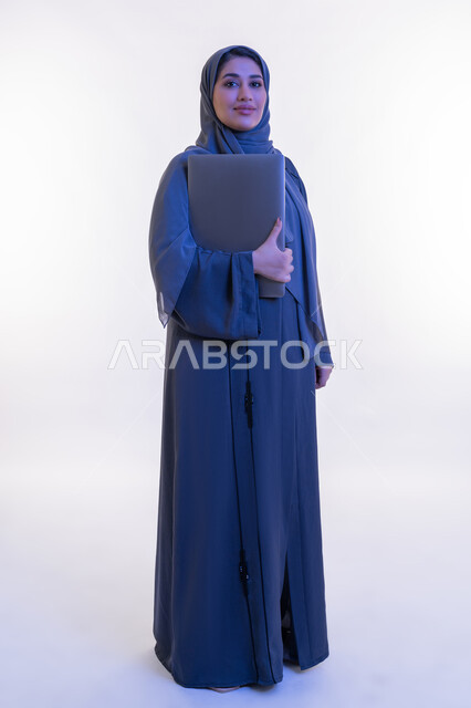 دمج التقنية والتكنولوجيا الحديثة في الحياة العملية، بورتريه لامرأة عربية خليجية اماراتية ترتدي العباءة تحمل بيدها جهاز الحاسوب المحمول، شابة سعودية تنظر الى الكاميرا بتعابير الثقة بالنفس والسرور، مهن ووظائف نسائية, صورة الجسم بالكامل، خلفية بيضاء