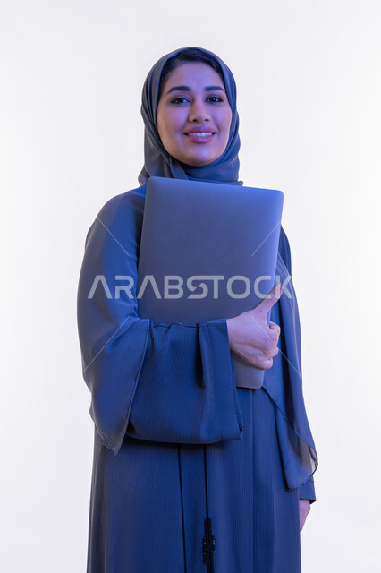 مهن ووظائف نسائية, دمج التقنية والتكنولوجيا الحديثة في الحياة العملية، بورتريه مقرب لامرأة عربية خليجية اماراتية ترتدي العباءة تحمل بيدها جهاز الحاسوب المحمول، شابة سعودية تنظر الى الكاميرا بتعابير الثقة بالنفس والسرور، مفهوم ادارة المشاريع عن بعد، خلفية بيضاء