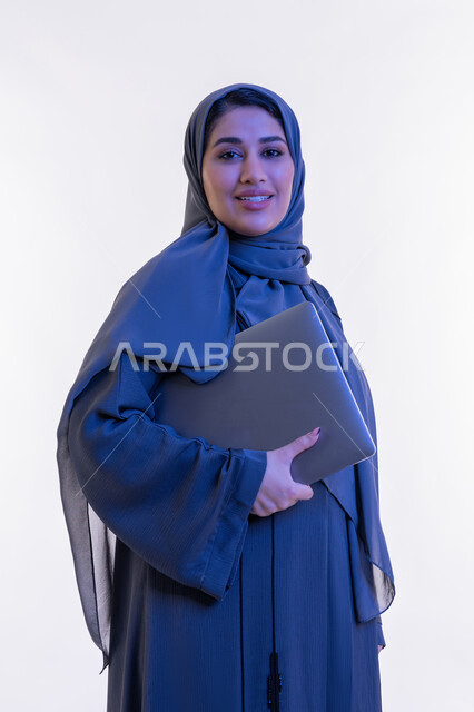مهن ووظائف نسائية, دمج التقنية والتكنولوجيا الحديثة في الحياة العملية، بورتريه مقرب لامرأة عربية خليجية اماراتية ترتدي العباءة تحمل بيدها جهاز الحاسوب المحمول، شابة سعودية تنظر الى الكاميرا بتعابير الثقة بالنفس والسرور، مفهوم ادارة المشاريع عن بعد، خلفية بيضاء