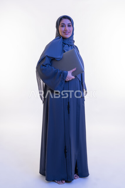 دمج التقنية والتكنولوجيا الحديثة في الحياة العملية، بورتريه لامرأة عربية خليجية اماراتية ترتدي العباءة تحمل بيدها جهاز الحاسوب المحمول، شابة سعودية تنظر الى الكاميرا بتعابير الثقة بالنفس والسرور، مهن ووظائف نسائية, صورة الجسم بالكامل، خلفية بيضاء