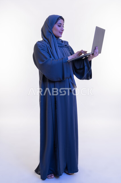 دمج التقنية والتكنولوجيا الحديثة في الحياة اليومية والعملية، بورتريه من الجانب لامرأة عربية خليجية اماراتية ترتدي العباءة تقوم بالنقر على لوحة مفاتيح اللابتوب، تصفح مواقع التواصل الاجتماعي، شابة سعودية تستخدم الحاسوب المحمول، الجسم الكامل بالطول، خلفية بيضاء