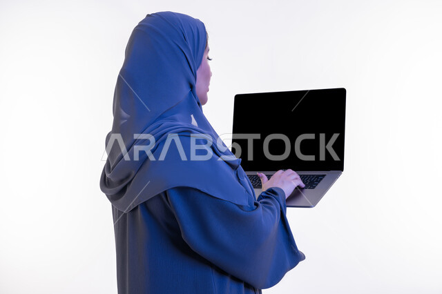 دمج التقنية والتكنولوجيا الحديثة في الحياة العملية، بورتريه مقرب من الخلف لامرأة عربية خليجية اماراتية ترتدي العباءة تقوم بالنقر على لوحة مفاتيح اللابتوب، شابة سعودية تعرض  شاشة الحاسوب المحمول سوداء، مفهوم ادارة المشاريع عن بعد، خلفية بيضاء