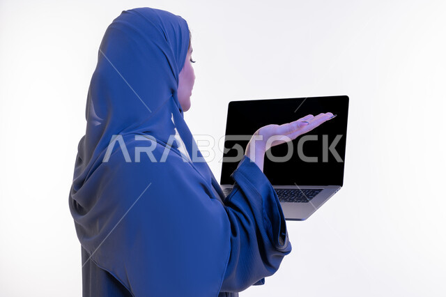 اجراء مقابلات عمل اونلاين، دمج التقنية والتكنولوجيا الحديثة في الحياة العملية، بورتريه مقرب من الخلف لامرأة عربية خليجية اماراتية ترتدي العباءة تحمل بيدها عنصر ما وترفع كف اليد بإيماءات عدم المعرفة، شابة سعودية تعرض شاشة الحاسوب المحمول سوداء، خلفية بيضاء