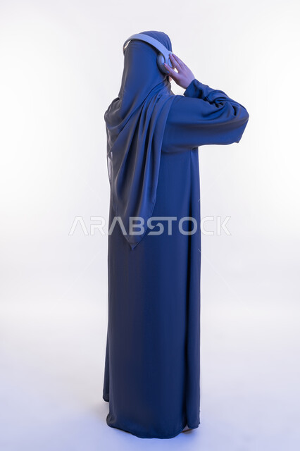Listening concept, having a good time on the weekend, a young Saudi woman using headphones, portrait from the back of a veiled Arab Gulf Emirati woman wearing an abaya wearing headphones listening to music, full body length, white background
