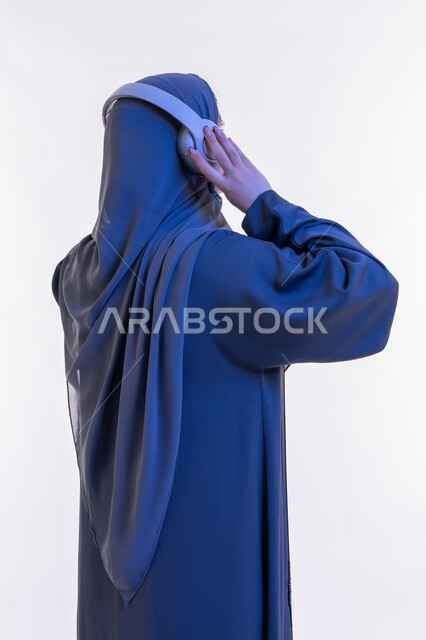 قضاء اوقات ممتعة في عطلة نهاية الاسبوع، شابة سعودية تستخدم  تقنية الهيدفون، بورتريه مقرب من الخلف لسيدة عربية خليجية اماراتية محجبة ترتدي العباءة تضع سماعات الرأس الصوتية تستمع إلى الموسيقى، مفهوم الإصغاء والإستماع، خلفية بيضاء