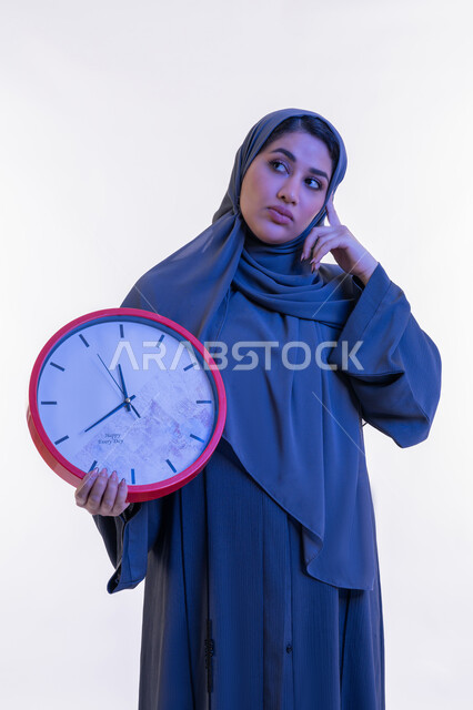 Importance of keeping time, completing tasks on time, close-up portrait of an Arab Gulf Emirati woman wearing an abaya holding a red wall clock, a young Saudi woman putting her hand on her head with expressions of thinking and contemplation, looking at a place with gestures reminding something, white background