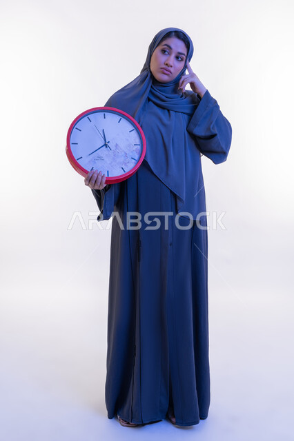النظر الى مكان ما بإيماءات تذكير شيء ما، أهمية الحفاظ على الأوقات، إنجاز المهام في الموعد المحدد, بورتريه لامرأة عربية خليجية إماراتية ترتدي العباءة تمسك بيدها ساعة حائط حمراء، شابة سعودية تضع يدها على رأسها بتعابير التفكير، الجسم الكامل بالطول، خلفية بيضاء