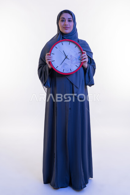 Importance of keeping time, completing tasks on time, constant reminders and alerts, portrait of an Arab Gulf Emirati woman wearing an abaya holding a red wall clock, young Saudi woman looking at the camera with gestures of pride and pleasure, full body length, white background